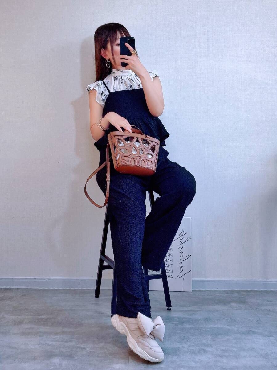 花柄ブラウスを着用した女性が黒いオールインワンを着て椅子に座りスマホを見ている