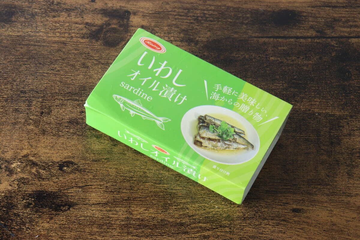 緑色のパッケージの「いわしオイル漬け」缶詰が斜め上から撮影され、木目のテーブルに置かれている 緑色のパッケージの「いわしオイル漬け」缶詰が斜め上から撮影され、木目のテーブルに置かれている