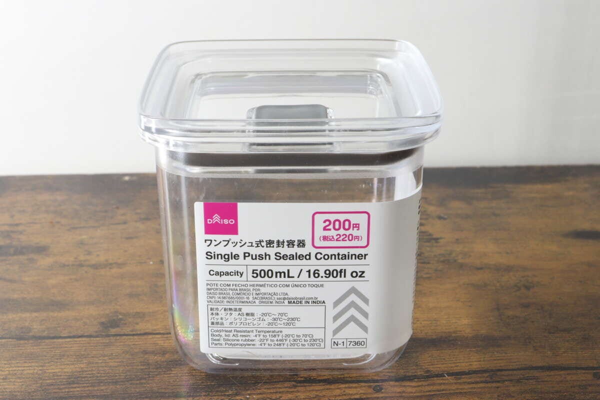 透明なワンプッシュ式密閉容器が木目のテーブルに置かれ、商品情報ラベルが貼られている 透明なワンプッシュ式密閉容器が木目のテーブルに置かれ、商品情報ラベルが貼られている