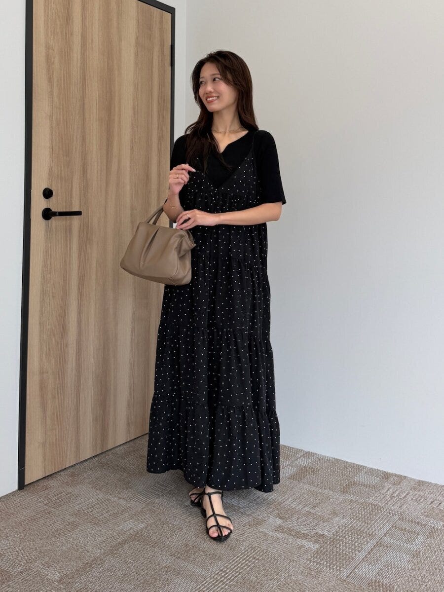 ドット柄の黒キャミワンピに黒ニットを重ね、ベージュのバッグを持つ女性が笑顔で立っている