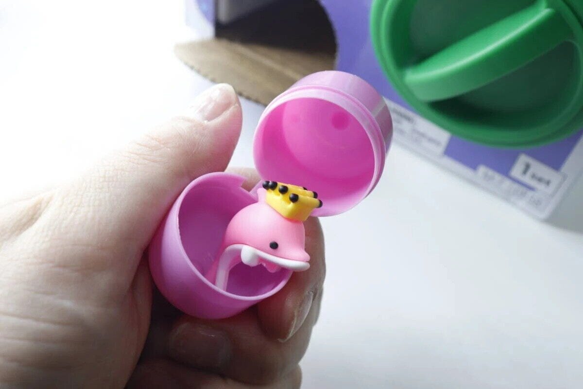 手がピンクのカプセルを開け、イルカのおもちゃが見える