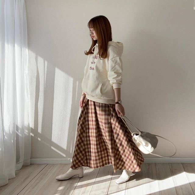 白のロゴ入りパーカーにブラウンのチェック柄フレアスカートを合わせ、白のスクエアブーツと白の巾着ショルダーバッグをコーデ