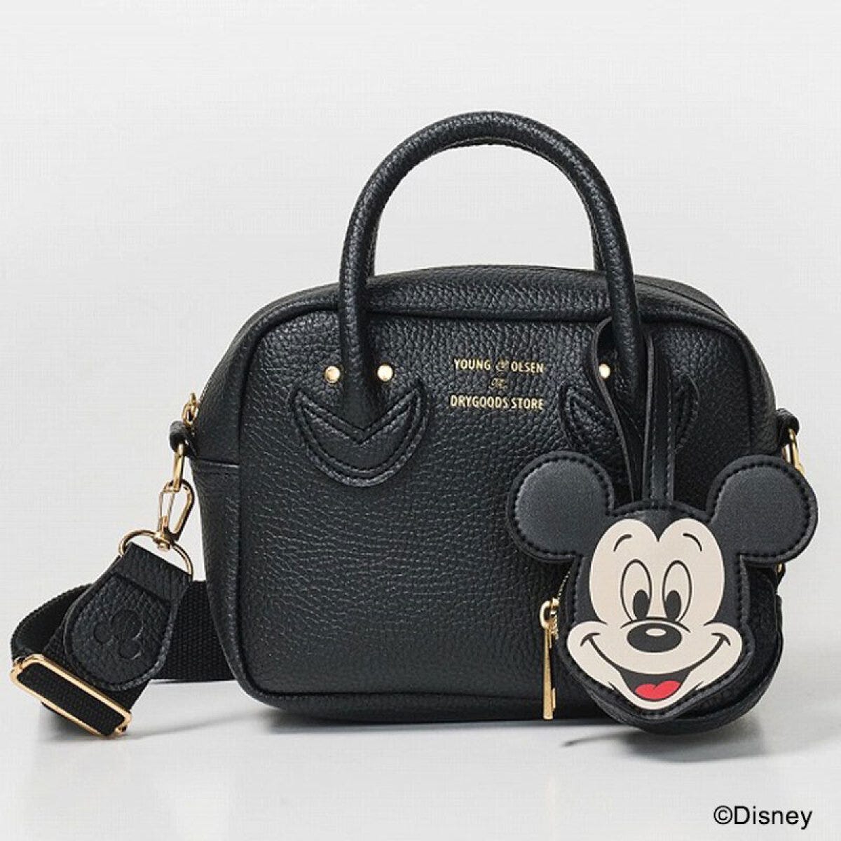 ヤングアンドオルセン×Disneyミッキーマウスフェイスチャーム付きショルダーバッグ 白背景にBLACKのシボ革風ボストン型ショルダーバッグ、サイドにミッキーマウスのフェイス型チャームポーチが下げられ、ストラップが垂れている斜め写真