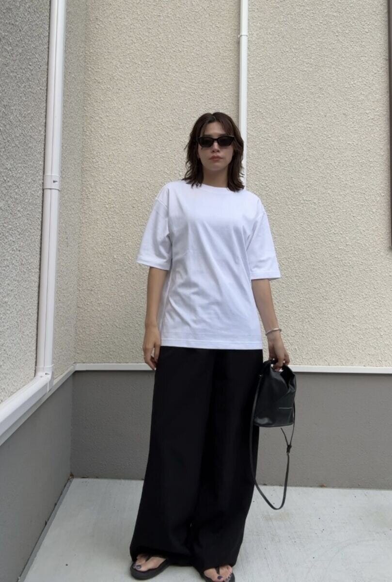 白いTシャツと黒いワイドパンツを着用し、サングラスをかけた女性がバッグを持って立っている