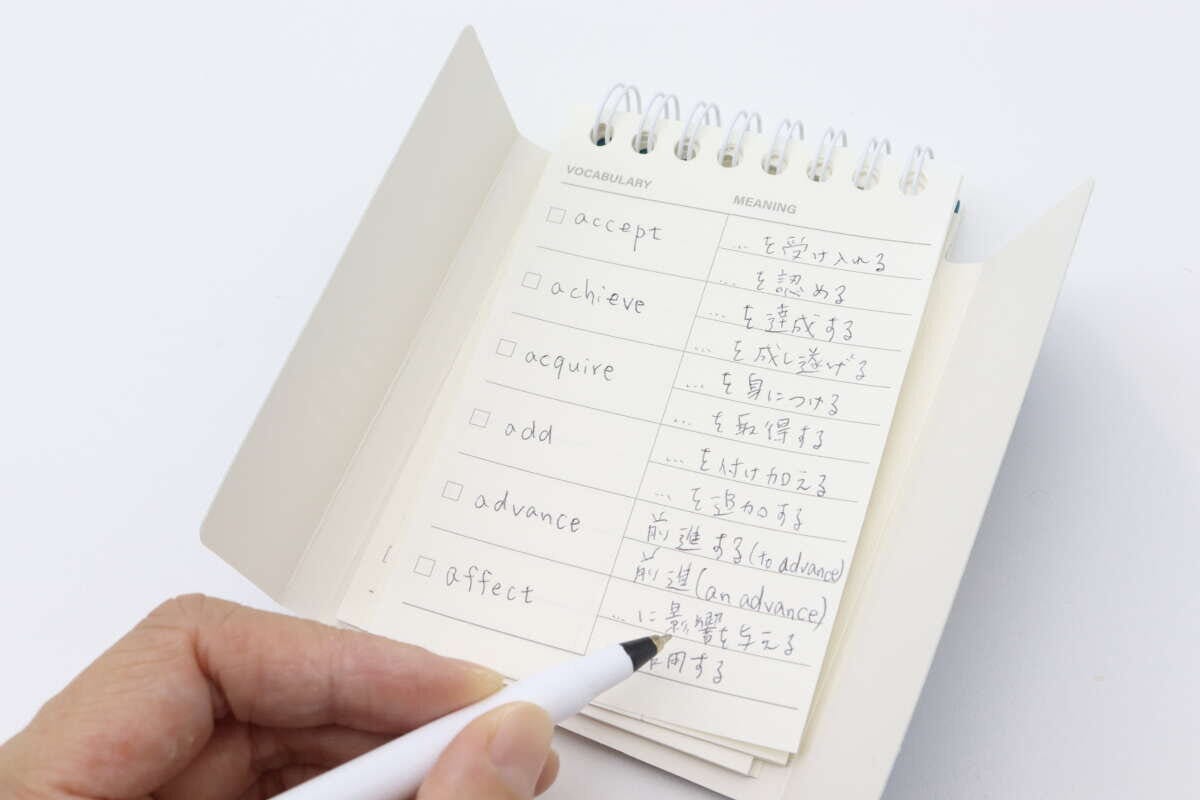 白いペンを持った手が、単語帳のページに英単語と意味を書き込んでいる様子 白いペンを持った手が、単語帳のページに英単語と意味を書き込んでいる様子