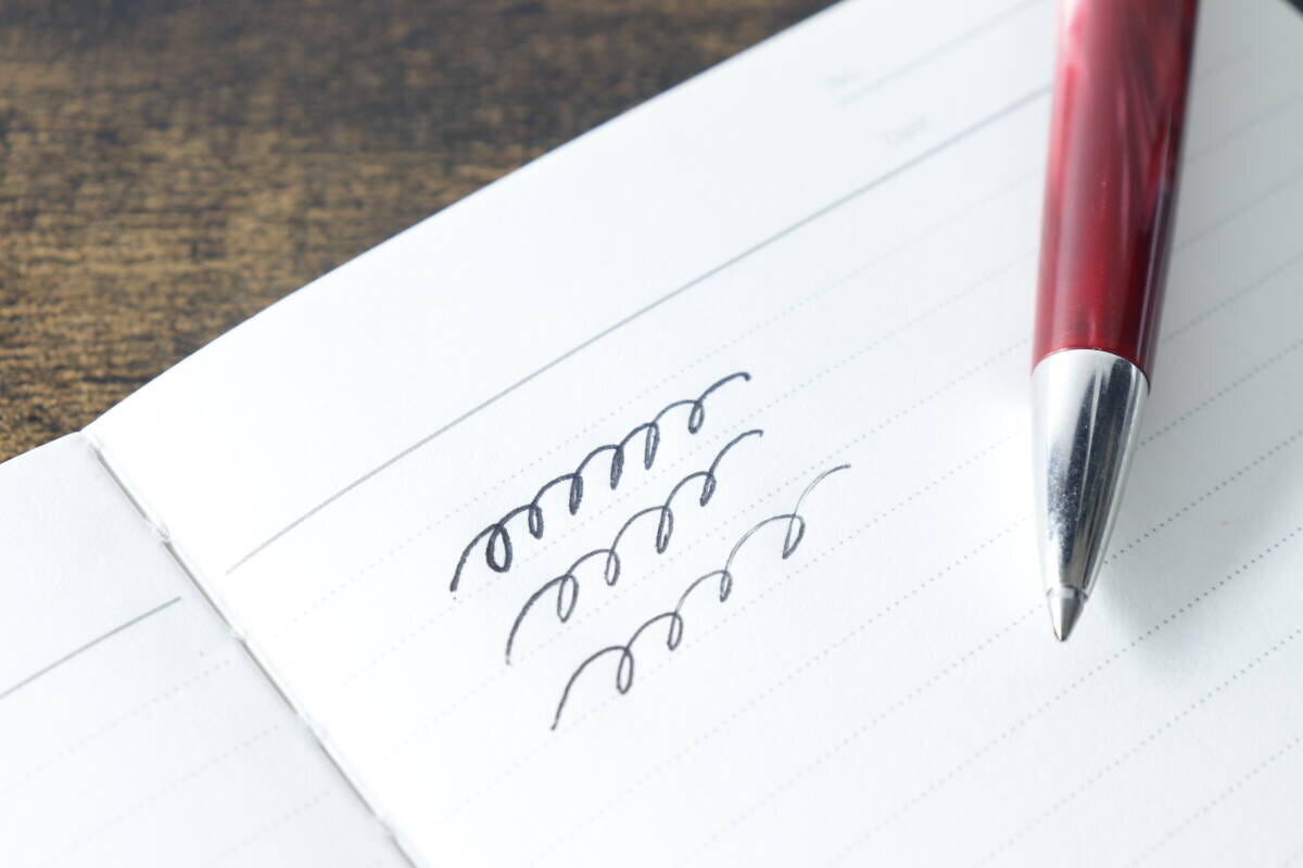ムーのレザー調ノートに赤いボールペンで文字を試し書きしている手元のクローズアップ ムーのレザー調ノートに赤いボールペンで文字を試し書きしている手元のクローズアップ