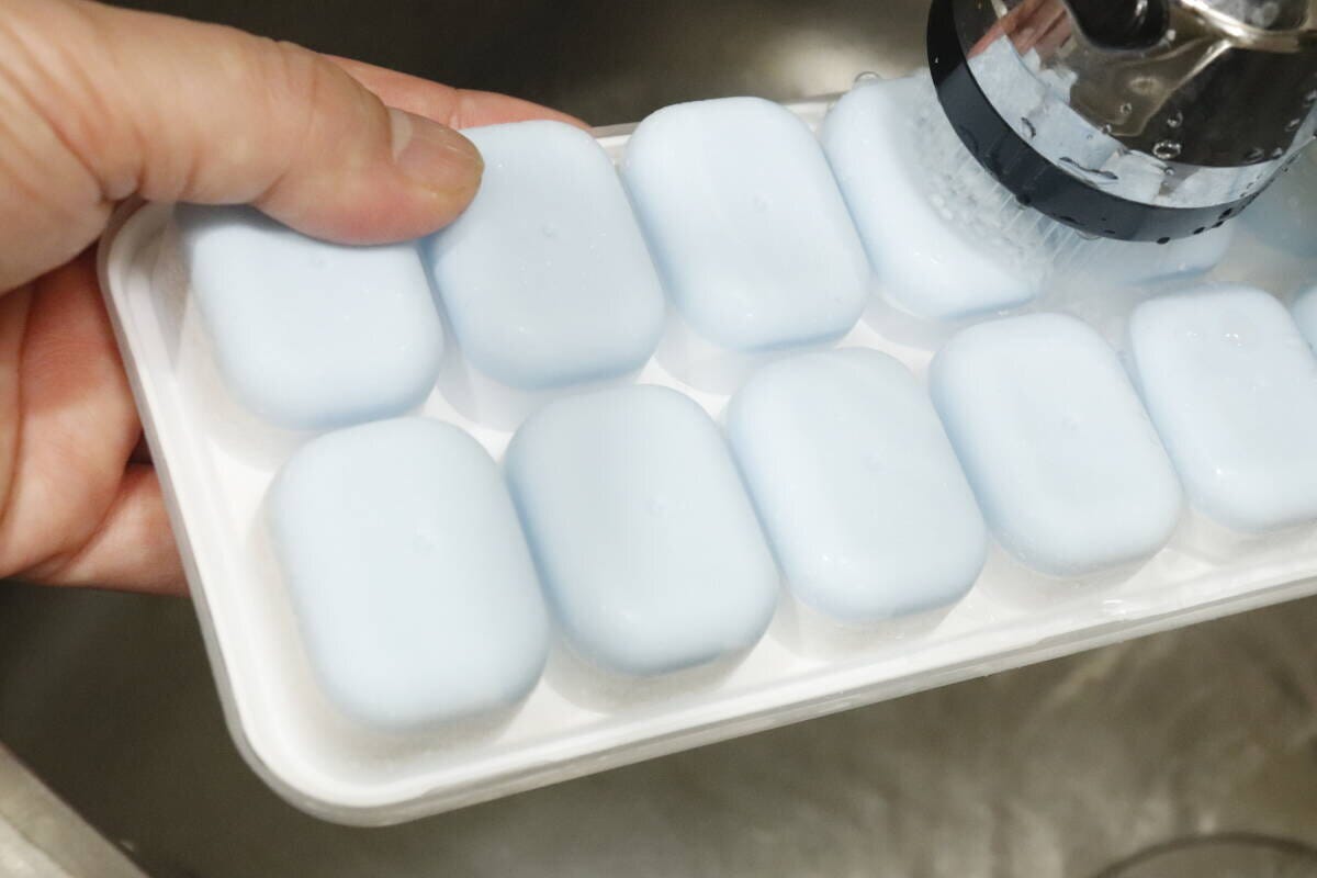 凍った氷が入ったアイストレーの底に、蛇口から出るシャワーで水をかけている手のアップ 凍った氷が入ったアイストレーの底に、蛇口から出るシャワーで水をかけている手のアップ