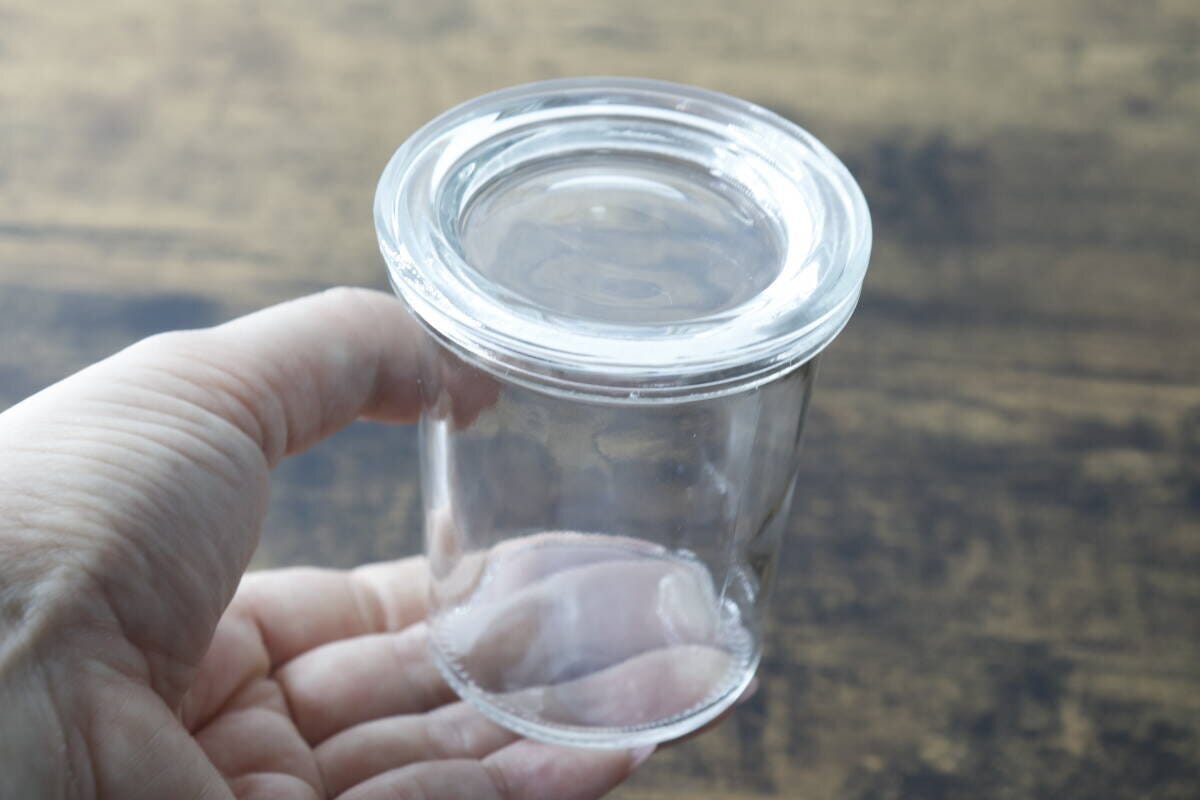 手のひらに乗せられた透明なガラス容器。蓋は外されており、容器の口が見える。 手のひらに乗せられた透明なガラス容器。蓋は外されており、容器の口が見える。
