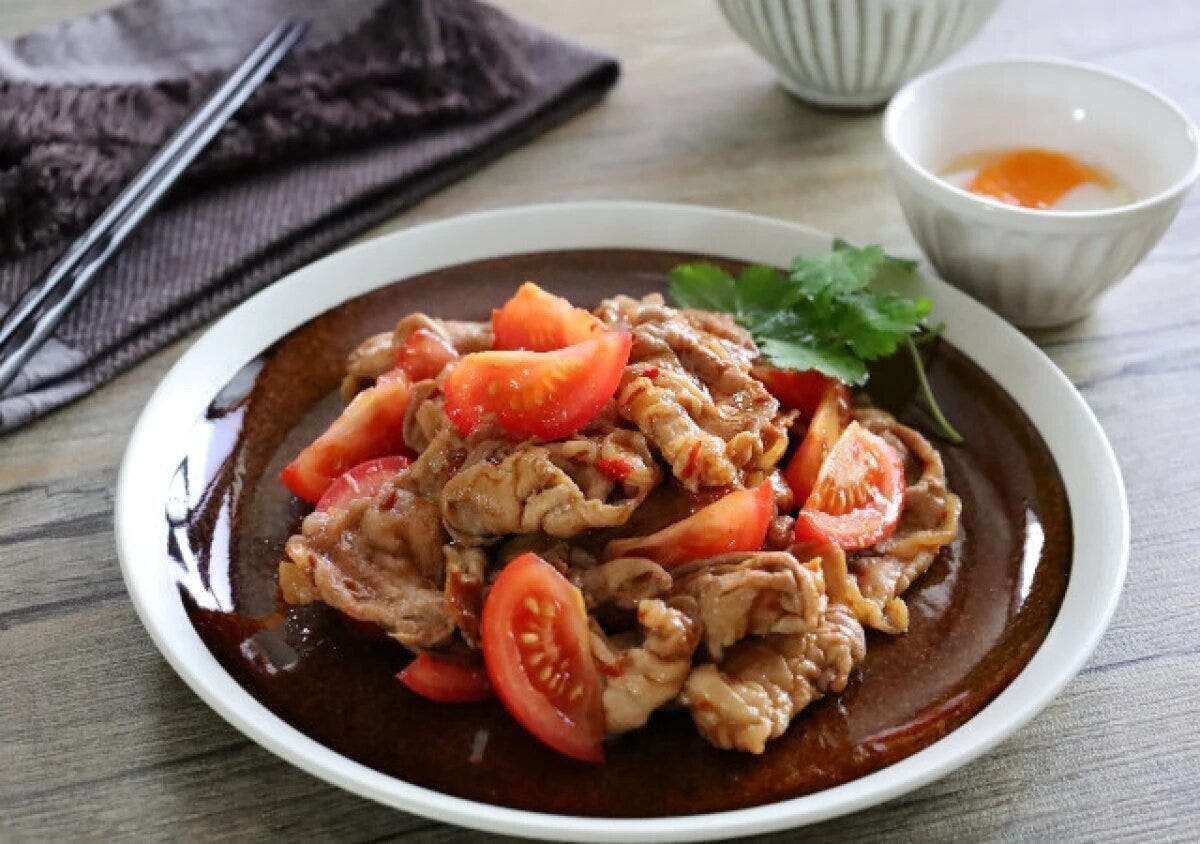 茶色い丸皿に盛られた、甘辛く炒めた薄切り豚肉と赤いくし切りトマト、添えられたパクチー。奥に温泉卵が入った白い小鉢