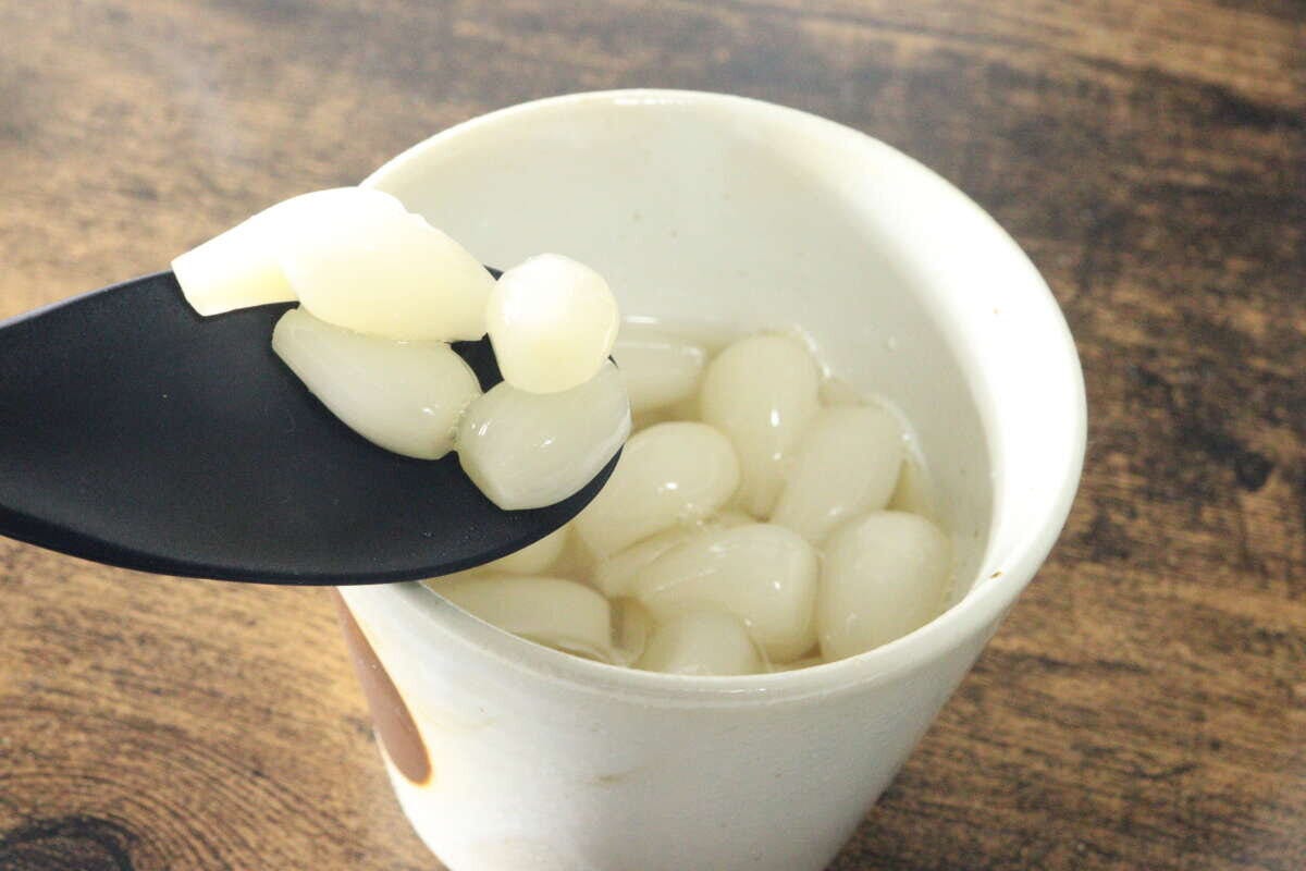 白い湯呑みに入った花らっきょうを黒いスプーンで数粒すくい上げている 白い湯呑みに入った花らっきょうを黒いスプーンで数粒すくい上げている