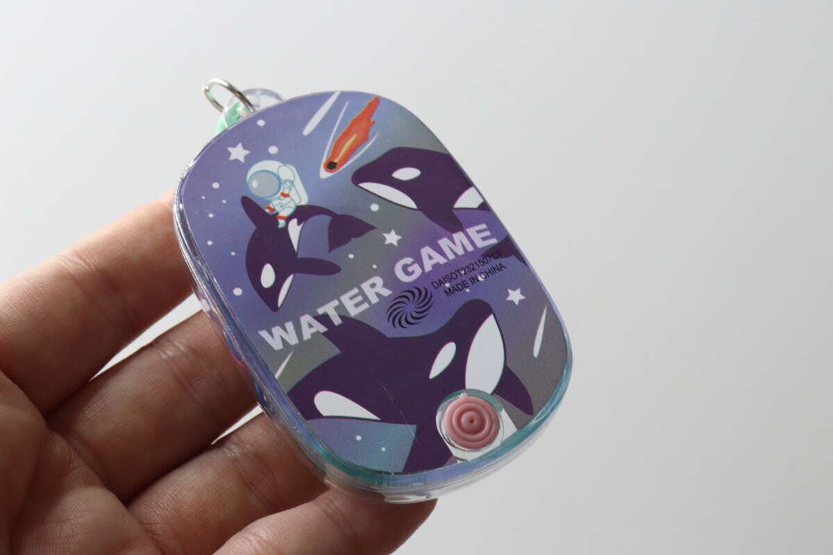 手のひらに乗せたミニウォーターゲーム機の裏側。宇宙服のシャチが描かれている 手のひらに乗せたミニウォーターゲーム機の裏側。宇宙服のシャチが描かれている