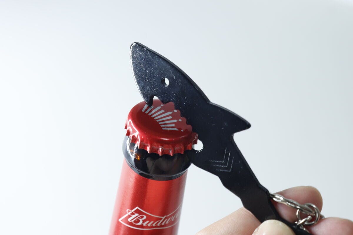 人の指が黒いサメ型栓抜きキーホルダーで赤い瓶ビールの蓋を開けようとしている