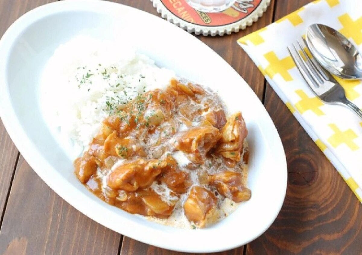 白い楕円形の皿に盛られたトマトバターチキンカレー。生クリームとパセリをトッピング。黄色いチェック柄のナプキンとカトラリーが添えられている