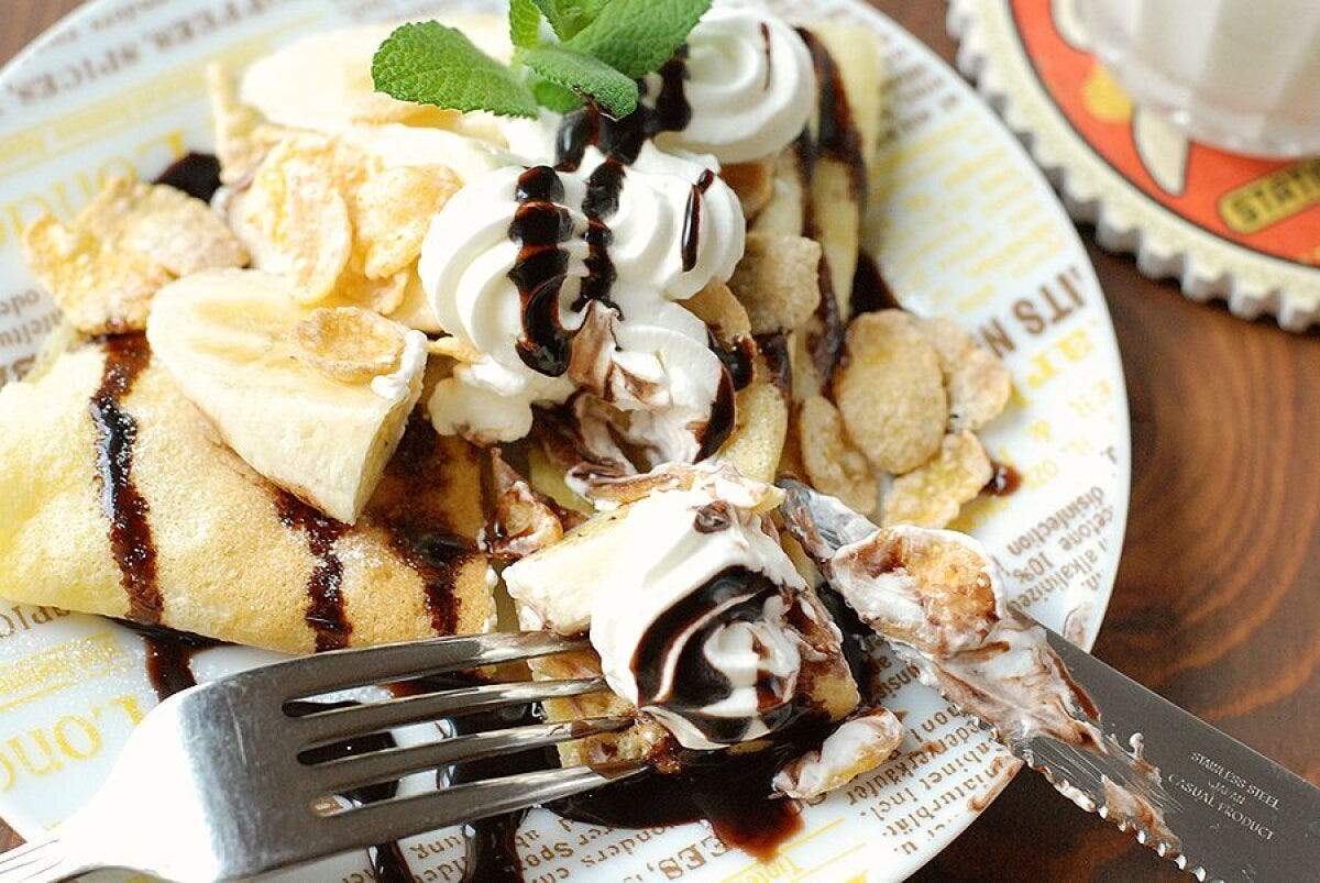 文字柄の皿に盛られた、ホイップクリームとスライスバナナ、チョコソース、コーンフレークをのせたクレープをフォークとナイフで切り分ける手元アップ
