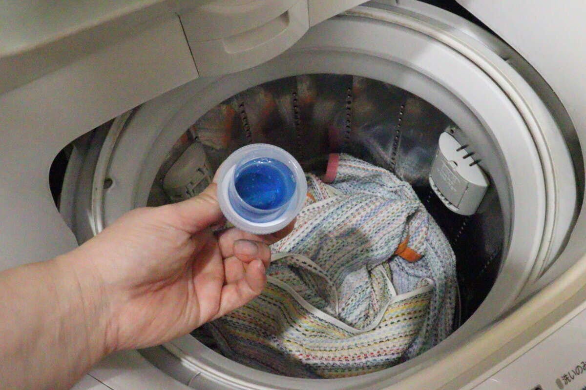 洗濯機の中に衣類が入っており、右手が計量カップに入った青い液体洗剤を注ごうとしている