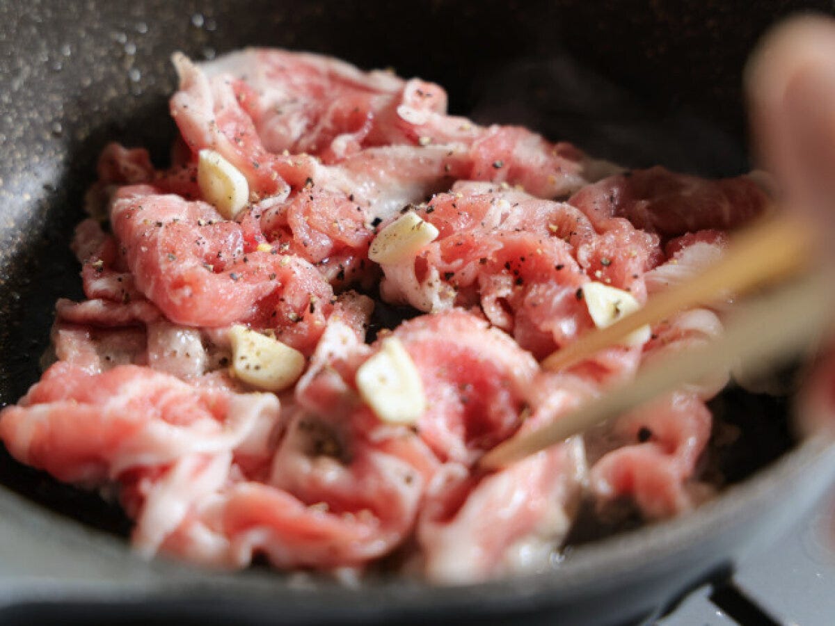 フライパンで薄切り豚肉と薄切りにんにくが塩こしょうで炒められており、箸で混ぜている様子。