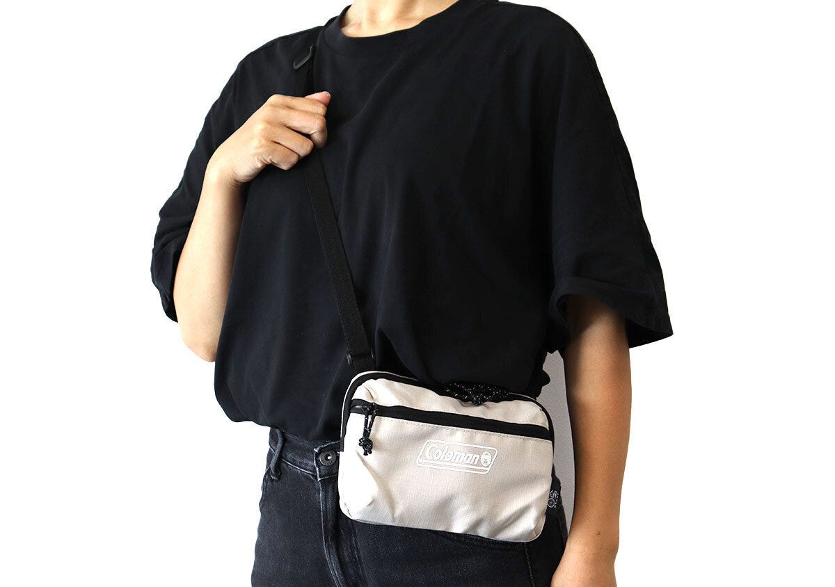 黒い半袖Tシャツの女性が右手でストラップを持ちColemanベージュのミニショルダーを斜め掛けする着用イメージ