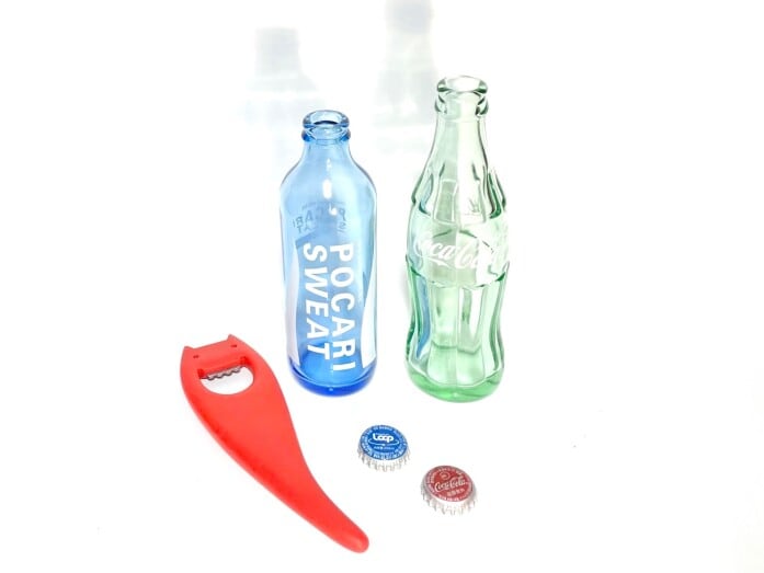 淡い色の付いたポカリとコーラの空瓶と王冠。コレクターの多い王冠のギザギザの数は世界的に21個で、この数は長年の研究の結果、落ち着いたと言われています。赤いオバケ形の栓抜きは1990年代に発売されたイタリア・アレッシィ社製のもので右にカーブした尻尾が握りやすく王冠があけやすい逸品