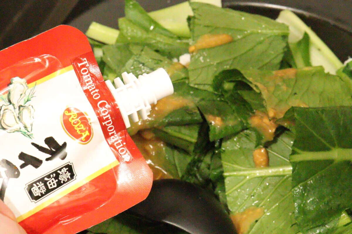フライパンで炒めている青菜に、ダイソーのオイスターソースのパウチからソースを注ぎ入れている手元