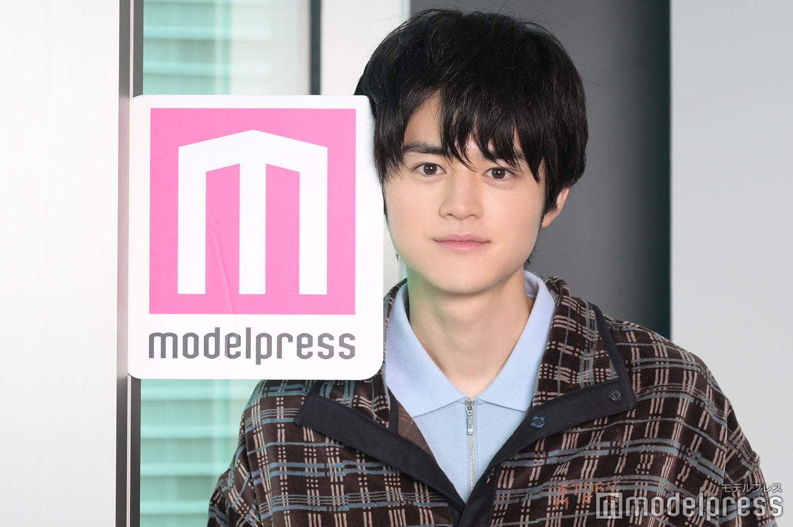 モデルプレスのロゴを壁と挟み遊んでいた鈴鹿央士さん（C）モデルプレス