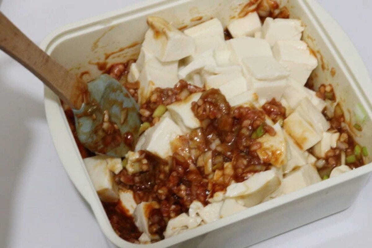 四角い容器の中で豆腐と麻婆豆腐の素を木製スプーンで混ぜている 四角い容器の中で豆腐と麻婆豆腐の素を木製スプーンで混ぜている