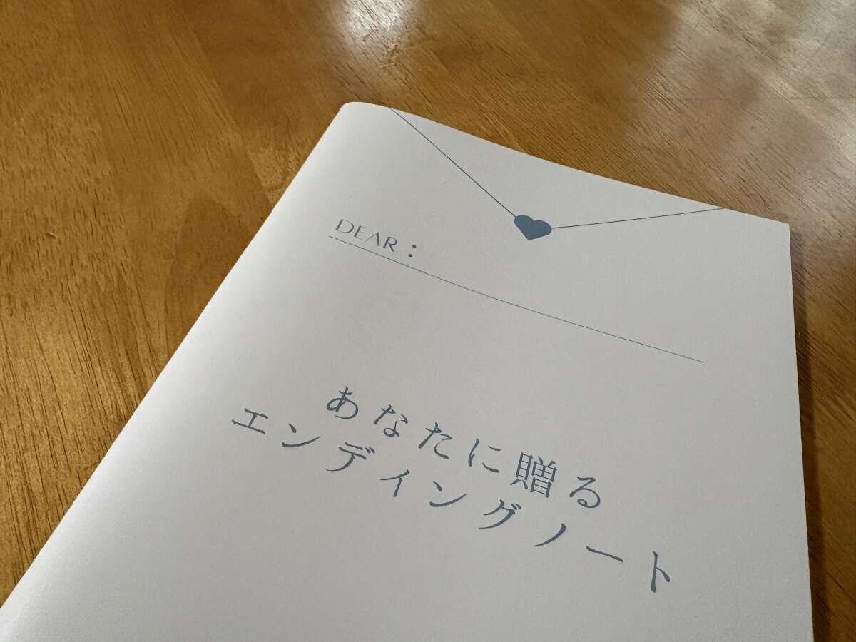 木目のテーブルに置かれた白い表紙の「あなたに贈るエンディングノート」。DEARの記入欄とハートマークが描かれている