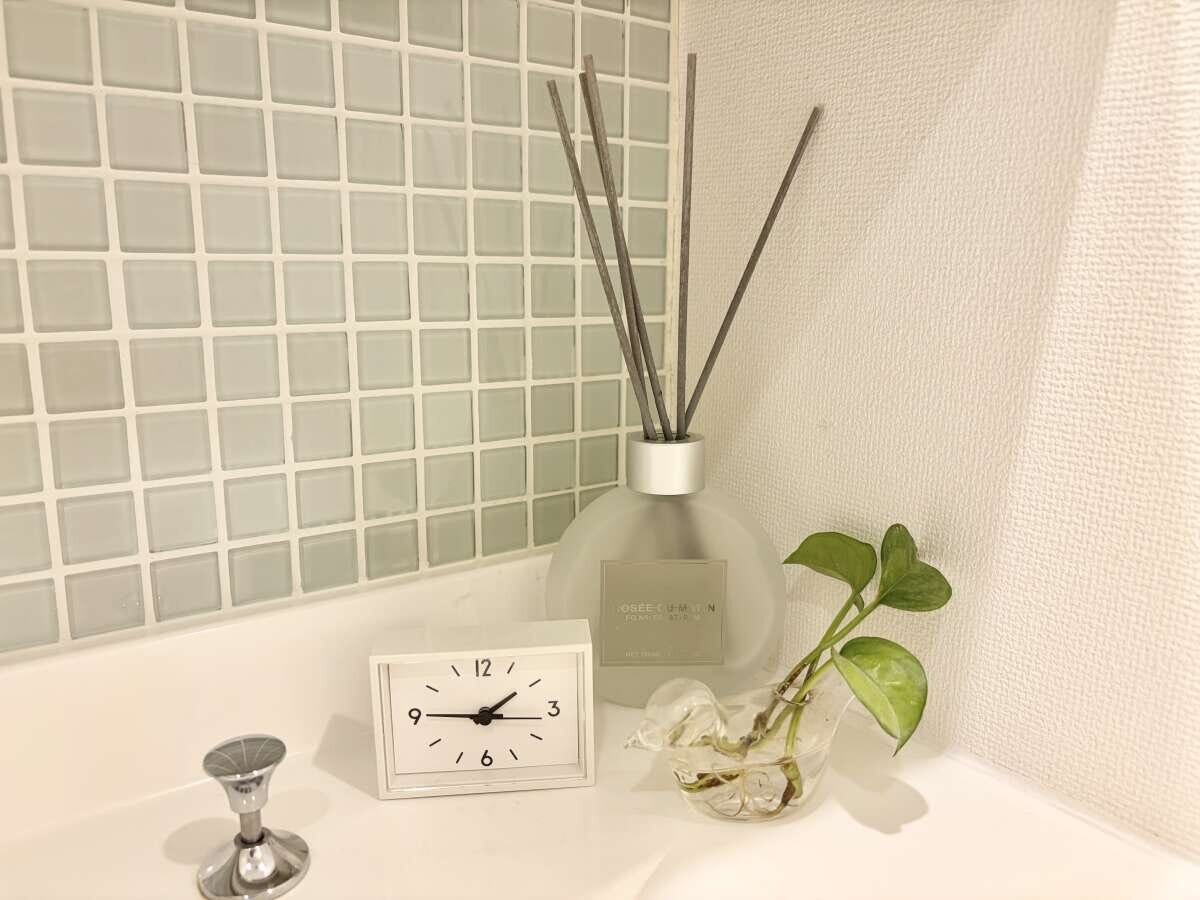 洗面台に飾った白い時計とディフューザーと観葉植物のディスプレイ グレーグリーンのタイルの洗面台にすりガラスのディフューザー・白いスクエア時計・水耕栽培のポトスが並んだ様子