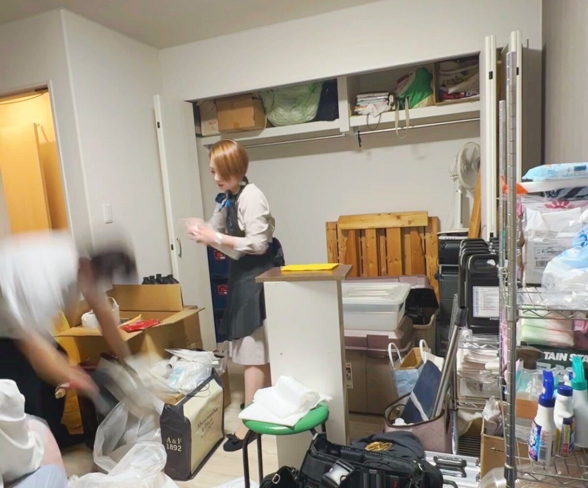 モノが多く散らかった部屋で、黒いエプロンを着けた整理収納アドバイザーがクローゼットの前で片付け作業をしている様子
