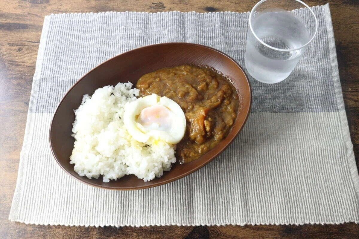 木目調オーバルプレートにカレーライスと温泉卵、グラスの水が食卓に置かれている