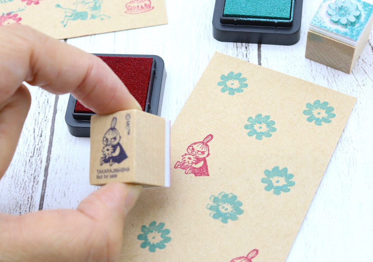 手がリトルミイのスタンプを持ち、クラフト紙に赤いインクで押している様子。紙には緑色の花柄スタンプも押されている。