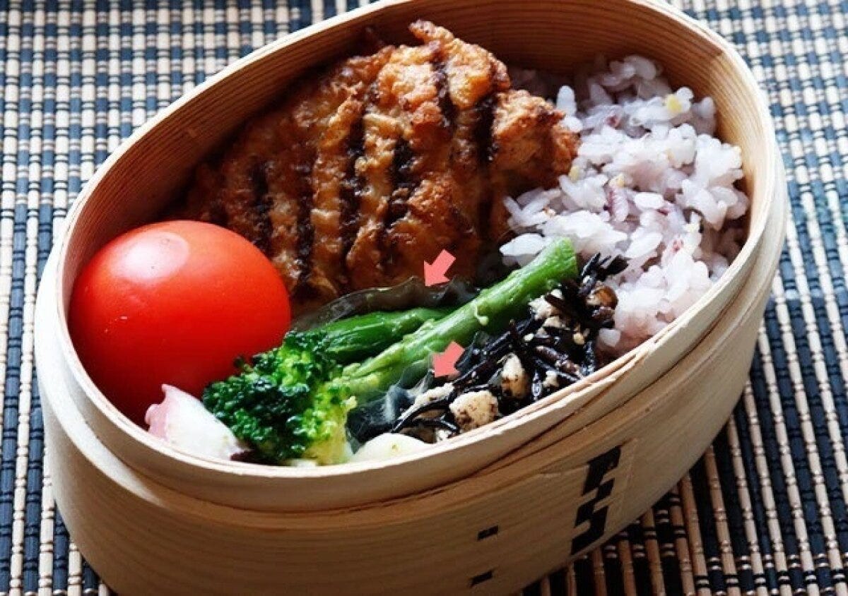 曲げわっぱに赤飯・唐揚げ・ブロッコリー・トマトなどを詰めた弁当に透明おかずカップを使った使用例、赤い矢印でカップの位置を示す